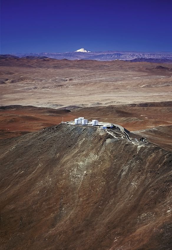 Photo aérienne de la demeure du Very Large Telescope (VLT) de l‘ESO sur le Mont Paranal au Chili. L‘observatoire de Paranal avec ses quatre télescopes géant de 8,2 mètres se trouve à une altitude de 2600 mètres, en arrière-plan, on peut voir le sommet enneigé de 6720 mètres d‘altitude du volcan Llullaillaco, situé à une distance de 190 km.