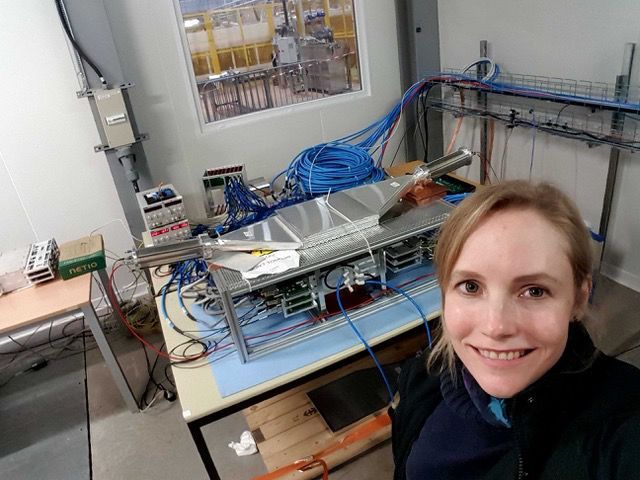 Claire Antel from the University of Geneva in front of the FASER cosmic ray test setup in an experimental hall at CERN above ground. She and her team use the measurement of cosmic rays from the atmosphere to test the triggering and data acquisition pipeline of FASER hardware and software components. The setup shown consists of 3 tracker planes sandwiched between 3 scintillators (which look like shiny paddles) — two on top and one on the bottom. The trigger system initiates the readout of data on the tracking planes when signals from all 3 scintillators arrive in coincidence, indicating the passage of a cosmic ray particle from above.